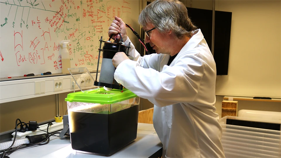 A man in a lab coat lowers an instrument into a small aquarium filled with dark liquid.
