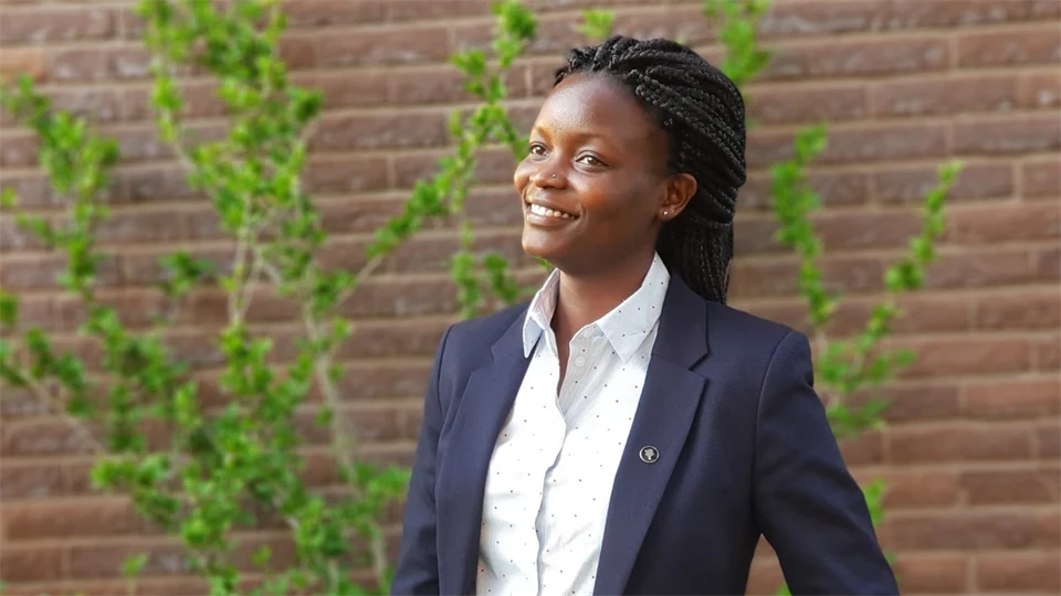 A woman with black hair up, blue jacket and white shirt standing by a brick wall.