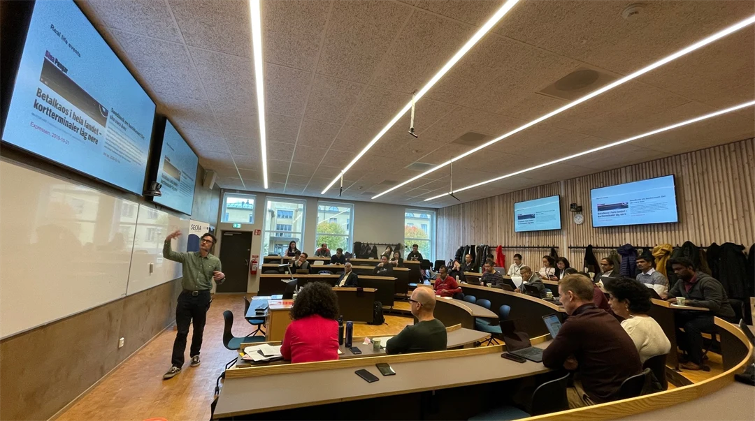 En sal med sittplatser i halvmåneform. Det sitter ett 30-tal personer i salen. Längst fram står en man och gestikulerar.