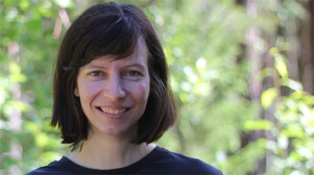 A woman with black hair and dark sweater stands outdoors and looks into the camera smiling.