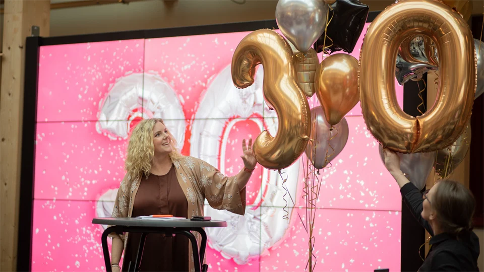 En kvinna står på en scen. Bredvid henne svävar ett antal ballonger i guld och silver. Två av ballongerna är formade som siffrorna 3 och 0. Hon tittar på en ballong och rör vid den.