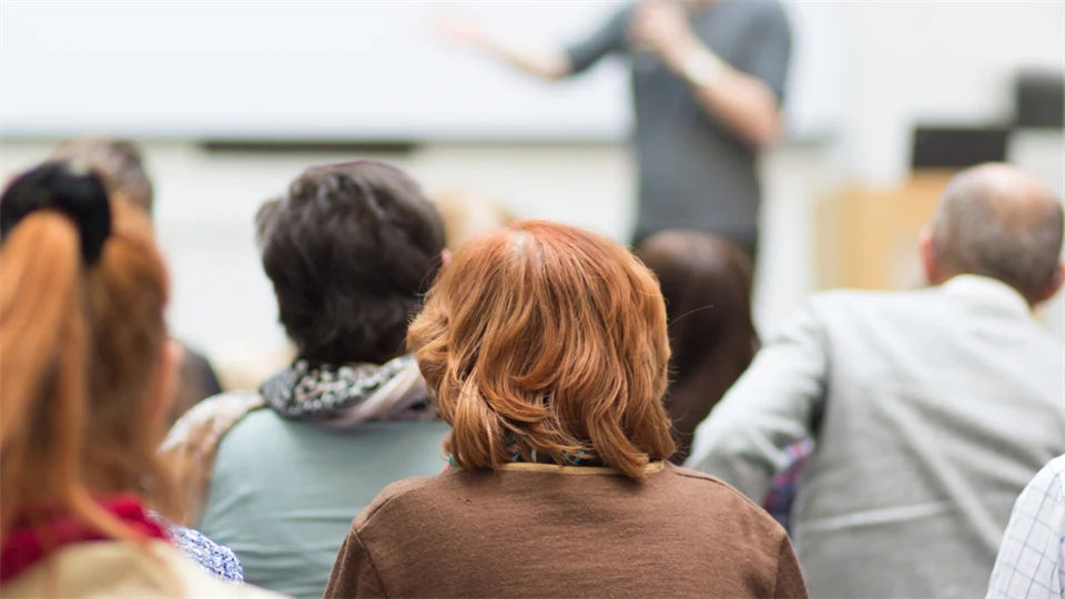 En man i bakgrund och ur fokus föreläser inför en sal där man endast ser fyggarna på åhörarna