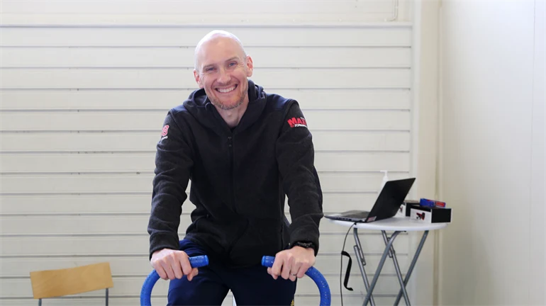 En leende mann sitter på en testcykel, bakom står ett bord med en bärbar dator.