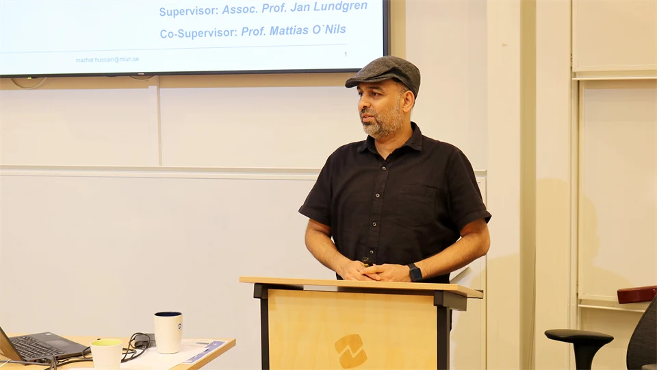 Man in black shirt and gray cap stands and gives a presentation.